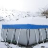Ein mit Schnee bedeckter, eckiger Stahlrahmenpool der mit einem Poolkissen, einer Abdeckplane und robusten Zurrgurten winterfest gemacht ist.