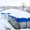 Ein mit Schnee bedeckter, eckiger Stahlrahmenpool der mit einem Poolkissen, einer Abdeckplane und robusten Zurrgurten winterfest gemacht ist.
